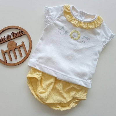 Conjunto de roupa de bebé branca e amarela com gola folhada e estampados, ao lado de objeto decorativo de madeira.