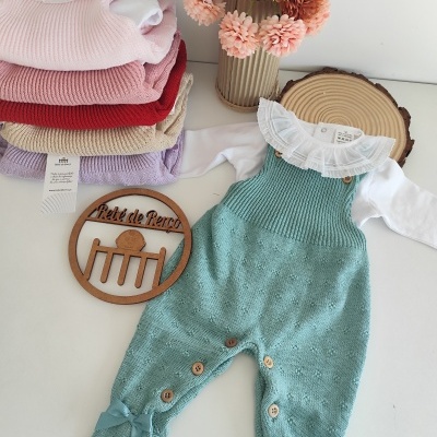 Conjunto de roupa de bebé com jardineira verde água e body branco junto a pilha de roupas coloridas e vaso com flores