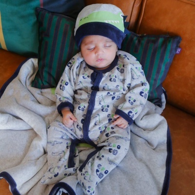 Bebé em macacão com estampado de robôs e chapéu, deitado em manta cinza e almofadas sobre sofá castanho