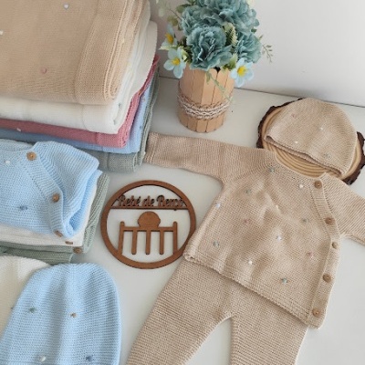 Conjunto de roupa de bebé em malha bege com botões e pontos coloridos, mantas empilhadas, chapéu, flores artificiais e placa de madeira com texto.