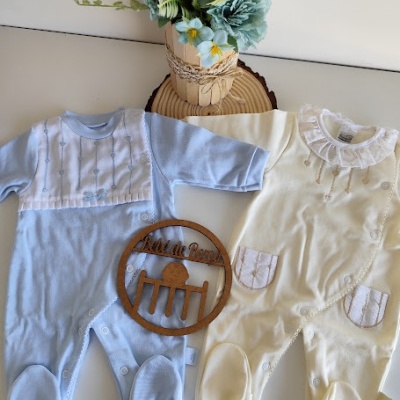 Dois macacões para bebé, um azul claro e outro amarelo claro, posados numa mesa com arranjo floral e placa de madeira