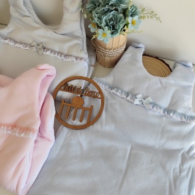 Sacos de dormir de bebé em cinza e rosa com rendas, flores artificiais e enfeite de madeira com texto Ateliê da Pinto