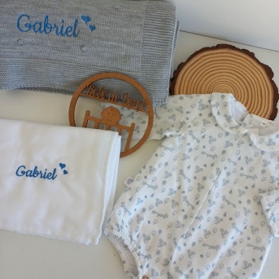 Conjunto de roupa para bebé com body, manta e pano branco com bordados azuis e objeto decorativo de madeira