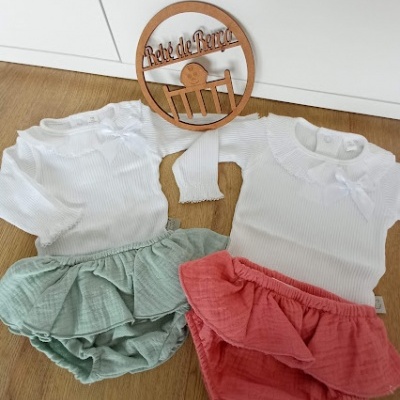 Dois conjuntos de roupa de bebé com saia verde e rosa e corpos brancos em chão de madeira