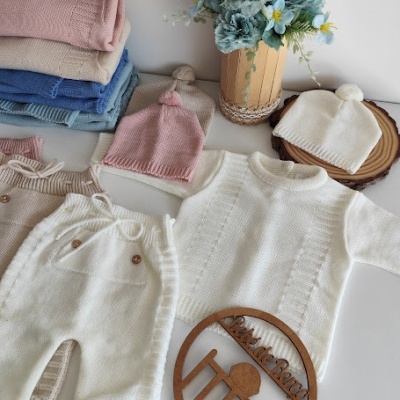 Conjunto de roupa de bebé em malha com camisolas, calças e gorros coloridos, ao lado de um vaso de flores azuis.