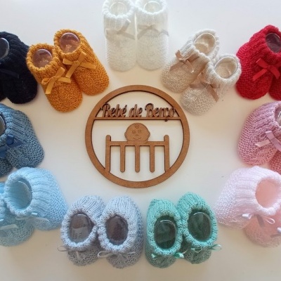 Sapatinhos de bebé de malha de várias cores dispostos em círculo sobre uma mesa branca com uma placa de madeira ao centro.