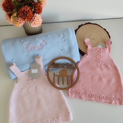 Dois vestidos de malha rosa claro, cobertor azul com nome bordado, vaso com flores secas e ornamento de madeira.