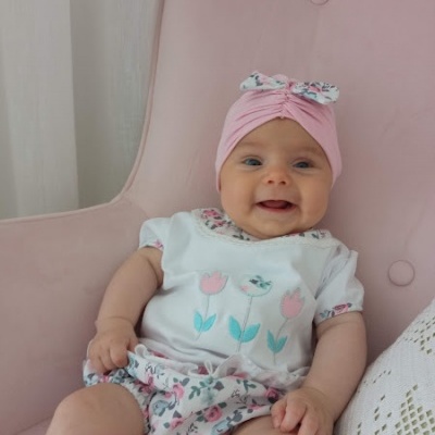 Bebé com roupa branca com flores, calções floridos e touca rosa sentada numa cadeirão cor de rosa.