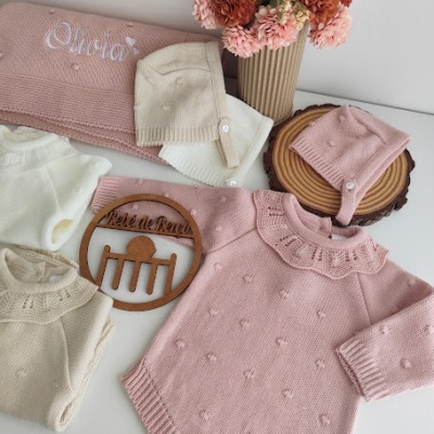Roupas de bebé em malha rosa, creme e branco com detalhes e um arranjo de flores