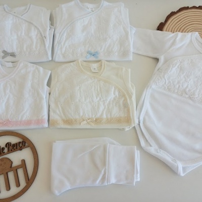Conjunto branco de roupa de bebé com detalhes em renda e laços coloridos sobre superfície branca e suporte de madeira com texto