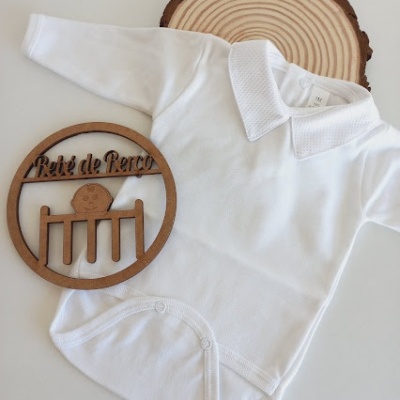 Body branco para bebé com gola e fechos de pressão, sobre base que imita madeira e placa de madeira decorativa.
