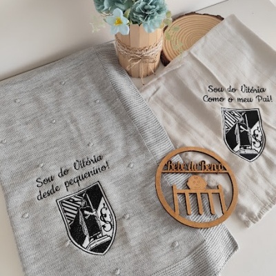 Tecidos bordados com brasão do Vitória, vaso de flores e emblema de madeira sobre superfície clara.