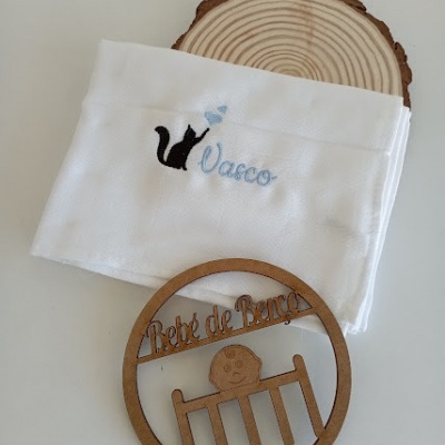 Toalha branca com bordado azul e preto e enfeite de madeira com texto 'Bebé de Berço'