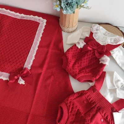 Conjunto de bebé vermelho com renda branca e laços, manta e roupa em malha, ao lado de vaso com flores azuis