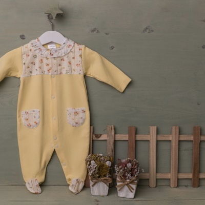 Babygrow amarelo com detalhes florais e botões, pendurado com cerca de madeira e vasos de flores secas ao lado