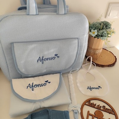 Conjunto de acessórios de bebé com mala azul claro, babete, cobertor, chupeta e camisola, com nome Afonso bordado, e placa em madeira com texto Retiró de Renê