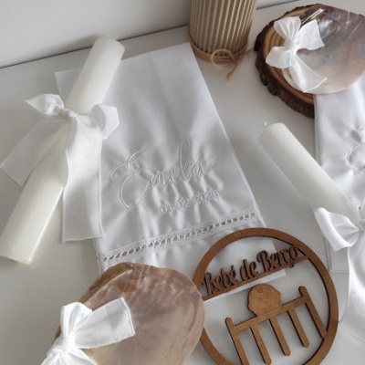 toalha bordada branca, vela branca com laço branco, conchas com laço branco, vaso com flores rosa, placa de madeira com texto