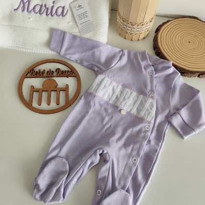 Macacão de bebé lilás com botões e faixa branca, toalha branca bordada com 'Maria' e decoração em madeira e flores