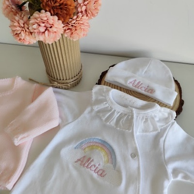 Roupinha de bebé branca com bordado 'Alicia' e flores artificiais num vaso