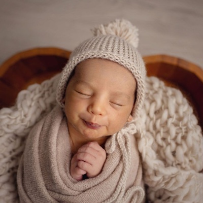 Bebé com gorro de malha bege e cobertor enrolado num cesto de madeira