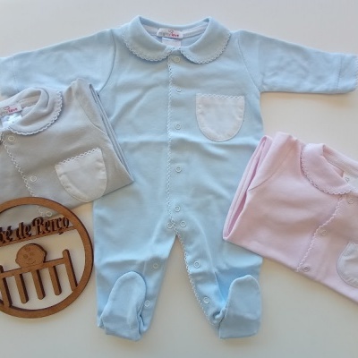 Três macacões de bebé nas cores azul, cinza e rosa com gola redonda e bolso frontal, junto a uma placa de madeira gravada.