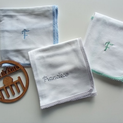 Três fraldas brancas com bordas coloridas e bordados, e uma placa redonda de madeira sobre fundo branco