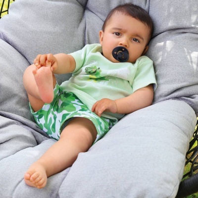 Bebé com roupa verde e calções camuflados numa almofada cinzenta com chupeta preta