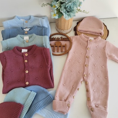 Roupa de bebé em malha colorida com textura de pequenas bolinhas e botões de madeira, incluindo macacão, casacos e gorros, e flores artificiais azuis ao fundo