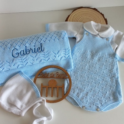 Conjunto de roupa de bebé em malha azul e branca com manta bordada e touca, placa de madeira e fundo branco