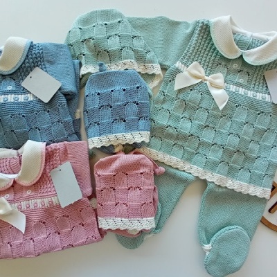 Conjuntos de roupa de bebé em tricô azul e rosa com golas brancas e laços, com etiquetas, sobre superfície clara e suporte de madeira