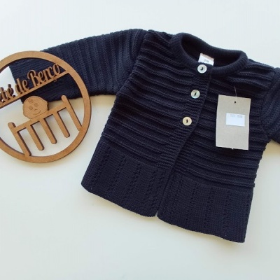 Cardigã azul-escuro de malha com botões, etiqueta pendurada, e objeto decorativo redondo de madeira com texto Arte de Barco