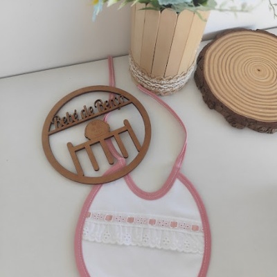 Babete de bebé branco com borda rosa e renda, enfeite de madeira redondo com texto e vaso de flores artificiais