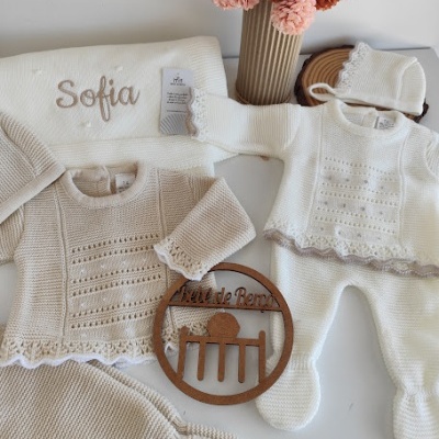Roupa de bebé em malha branca e bege com manta personalizada, flores em vaso e decoração de madeira
