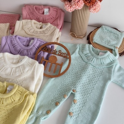 Roupas de bebé em malha colorida com botão de madeira e estampas, sobre mesa branca com flores e placa Bébé de Berço.