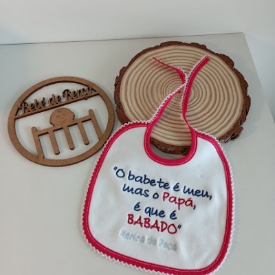 Babete branco com bordas vermelhas e texto bordado, sobre mesa branca com decorações de madeira redondas