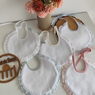 Cinco babetes de bebé com bordas de renda em várias cores e vaso com flores artificiais