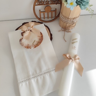 Toalha branca com renda, concha decorativa com laço rosa, vela branca com laço, vaso com flores azuis e placa de madeira
