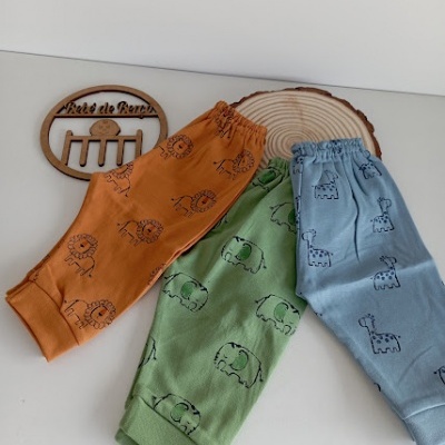 Três calças infantis coloridas com estampas de animais sobre superfície branca e objetos em madeira