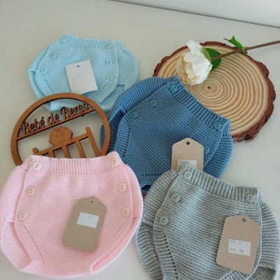 Calções de malha para bebés em azul claro, azul médio, rosa e cinzento com botões e etiquetas sobre superfície branca com flor e madeira redonda.