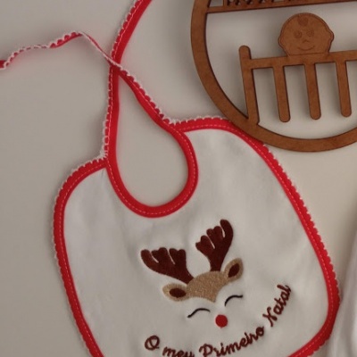 Babete branco e vermelho com rena bordada e texto, ao lado uma placa de madeira decorativa para bebé