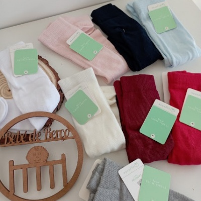 Meias coloridas para bebé com etiquetas verdes da marca 'Babybasics' sobre mesa branca, com decoração em madeira 'Bebé de Berço'