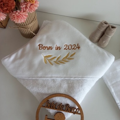 Toalha de banho branca com bordado beige 'Born in 2024', sapatos de bebé bege, flores cor de rosa e laranja, placa redonda de madeira 'Boîte de Bébé'