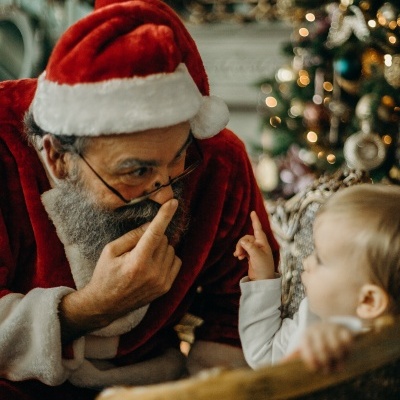 Pai Natal com barba e criança numa cadeira de vime junto a árvore de Natal decorada