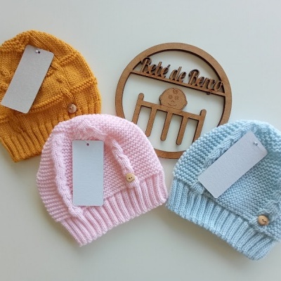 Três gorros de malha para bebé em mostarda, rosa e azul claro com etiquetas, botões de madeira e placa de madeira 'Pé de Benta'
