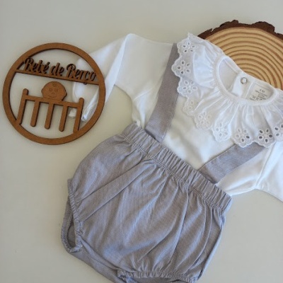 Conjunto de bebé branco com gola de renda e calções cinzentos com alças.