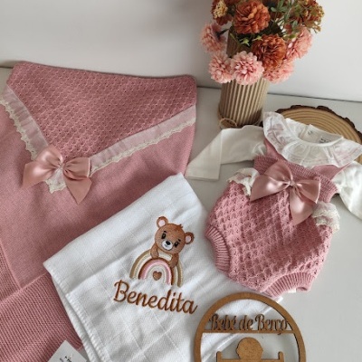 Conjunto de roupa de bebé rosa com laços, manta e toalha bordada, e placa decorativa em madeira com vaso de flores