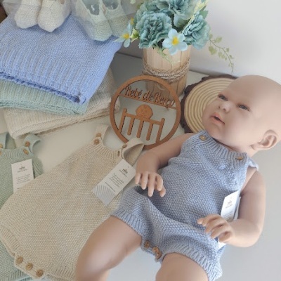 Boneco bebê com macacão azul claro de malha e sapatinhos, rodeado por macacões e sapatinhos de malha, manta azul, flores e placa decorativa em madeira