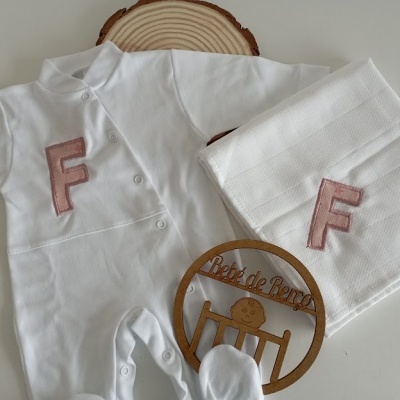 Conjunto branco de roupa de bebé com letra F rosa e manta em fundo branco com elementos decorativos em madeira