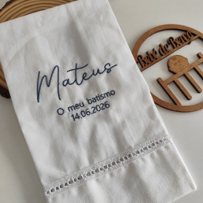 Toalha branca bordada para batismo com nome e data, sobre superfície branca com objetos decorativos de madeira