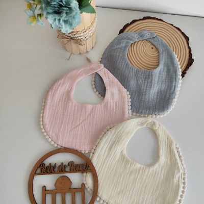 Três babetes de algodão cor cinza, rosa e creme com pompons, arranjo floral e ornamento de madeira